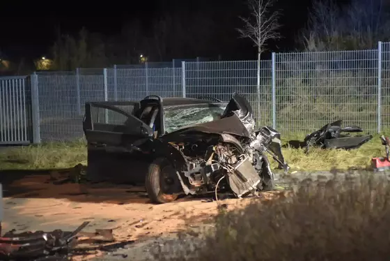 Schwer beschädigtes Auto mit aufgeschlagener Motorhaube und geöffneter Fahrertür vor einem Metallzaun bei Nacht.