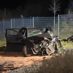 Schwer beschädigtes Auto mit aufgeschlagener Motorhaube und geöffneter Fahrertür vor einem Metallzaun bei Nacht.