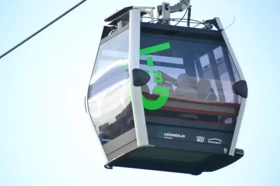 Seilbahnkabine mit grüner Aufschrift 'GrünBerlin Seilbahn' und Logos von 'Gärten der Welt' und 'Kienbergpark' an einer Seilbahnleitung hängend.