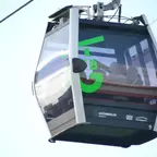 Seilbahnkabine mit grüner Aufschrift 'GrünBerlin Seilbahn' und Logos von 'Gärten der Welt' und 'Kienbergpark' an einer Seilbahnleitung hängend.