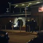 Polizisten vor einem Holzzaun bei Nacht, zwei öffnen eine Tür und leuchten mit Taschenlampen.