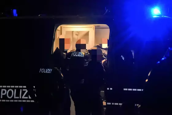 Polizisten laden bei Nacht eine Person in ein Fahrzeug mit geöffneter Tür und eingeschaltetem Blaulicht.